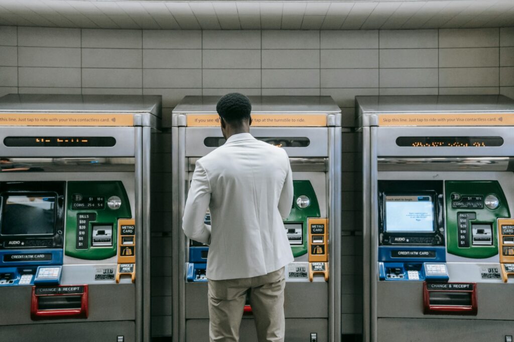 A black man in a white jacket and tan pants uses an ATM. There are empty ATMs to the left and right, creating a very symmetrical scene.