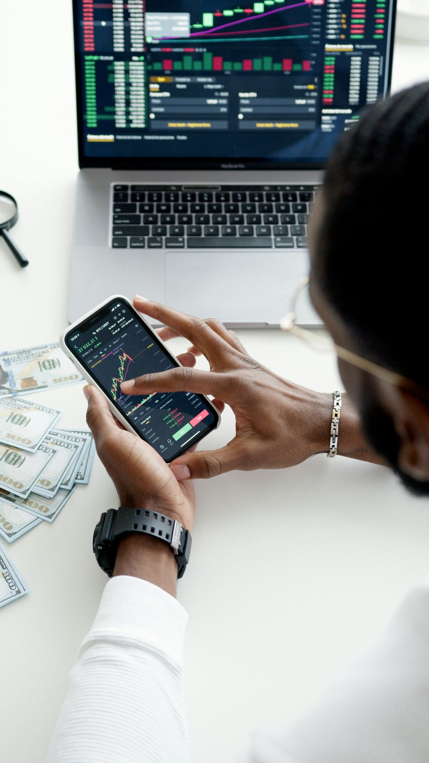 Photo is taking over the shoulder of a black man. They are looking at an investment app on their smart phone. In the background on the table is a pile of cash and a macbook open to an investment app.