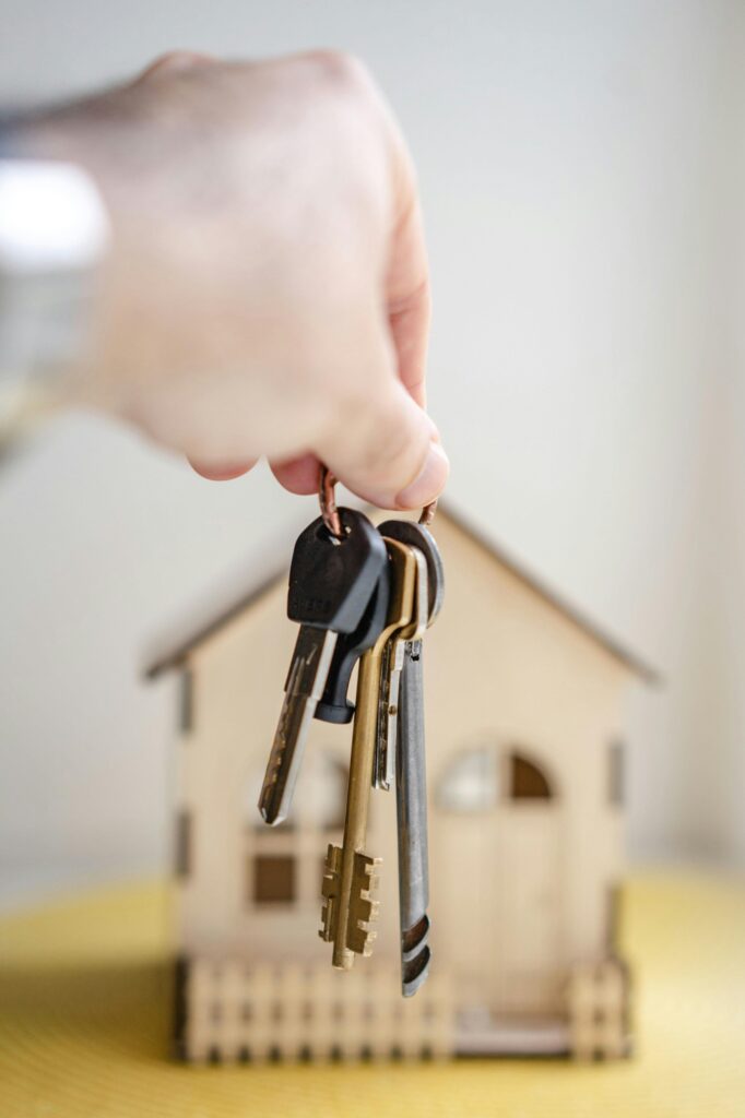 An arm holding up keys in front of a wooden miniture house.