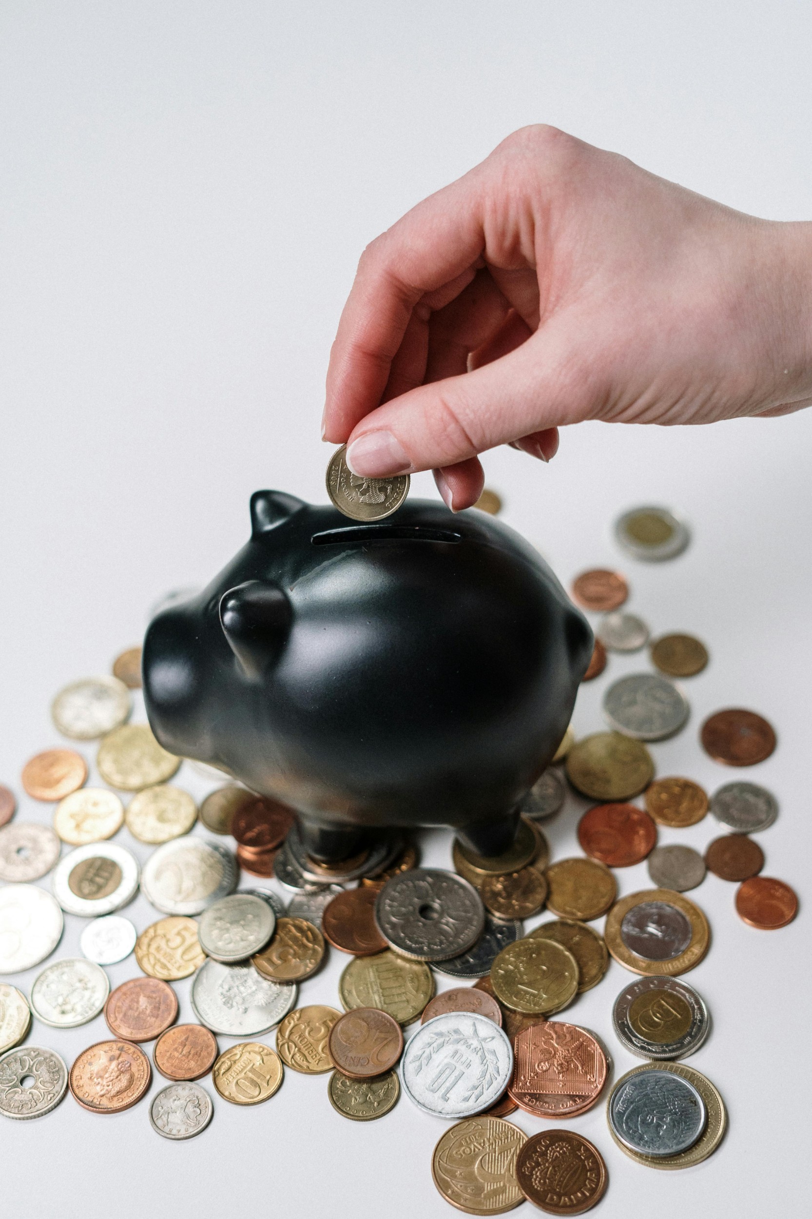 A hand putting money in a black piggy bank. There are coins scattered below the piggy bank.